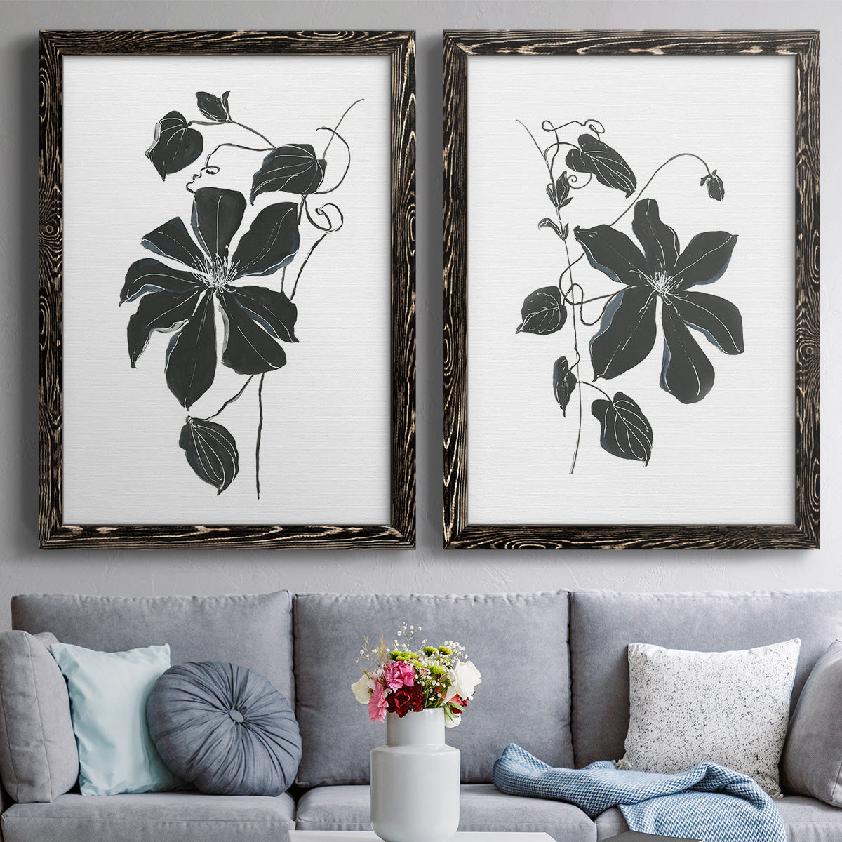 Set of two black barnwood framed canvas prints depicting paired minimalist botanical prints with bold dark leaves, curving stems, and clean white grounds above a sofa in a contemporary living room.
