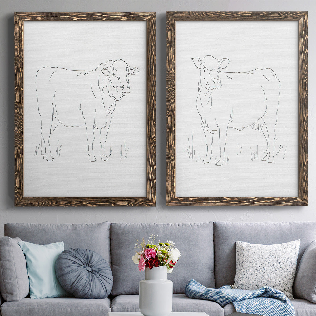 Set of two walnut barnwood framed canvas prints depicting paired minimalist livestock sketches with fine gray lines, white backgrounds, and quiet farm-animal profiles above a sofa in a contemporary living room.