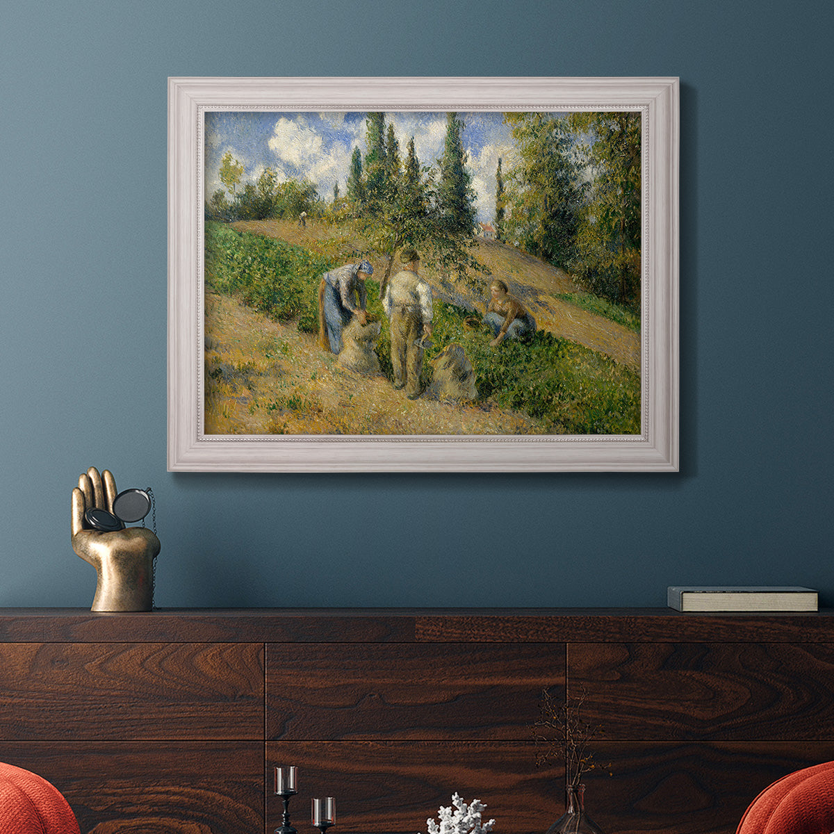 Impressionist countryside harvest scene by Camille Pissarro in an ornate weathered white frame displayed in a dark-toned interior setting.