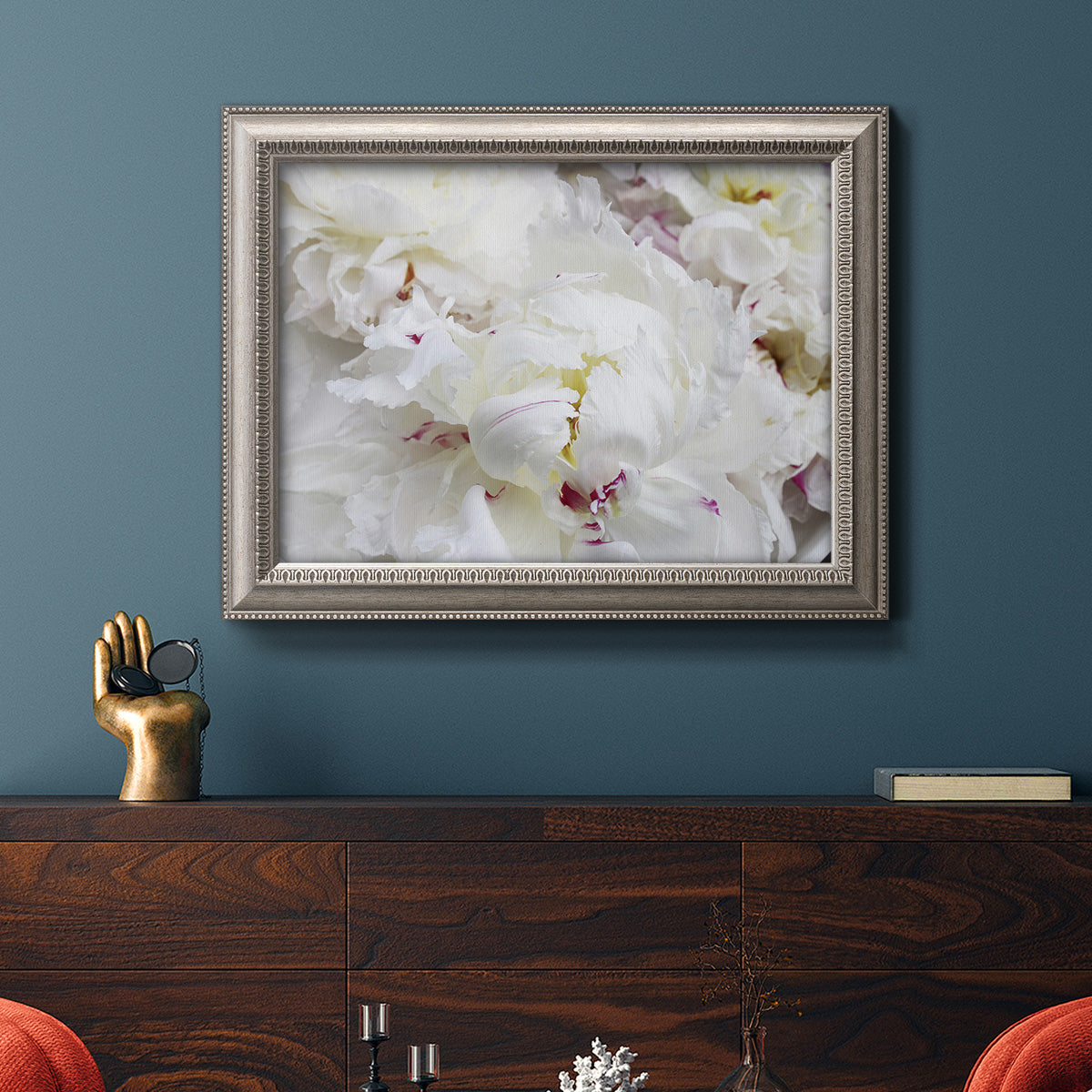 White peonies in bloom in an ornate vintage silver frame displayed in a dark-toned interior setting.