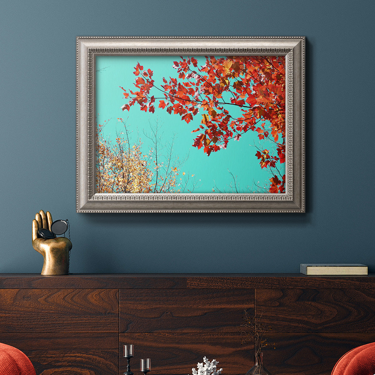 Red autumn leaves against a blue sky in an ornate vintage silver frame displayed in a dark-toned interior setting.