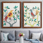 Set of two maple barnwood framed canvas prints depicting paired modern folk-art bird illustrations with red yellow blue plumage, simplified leaves, and playful flat shapes above a sofa in a contemporary living room.