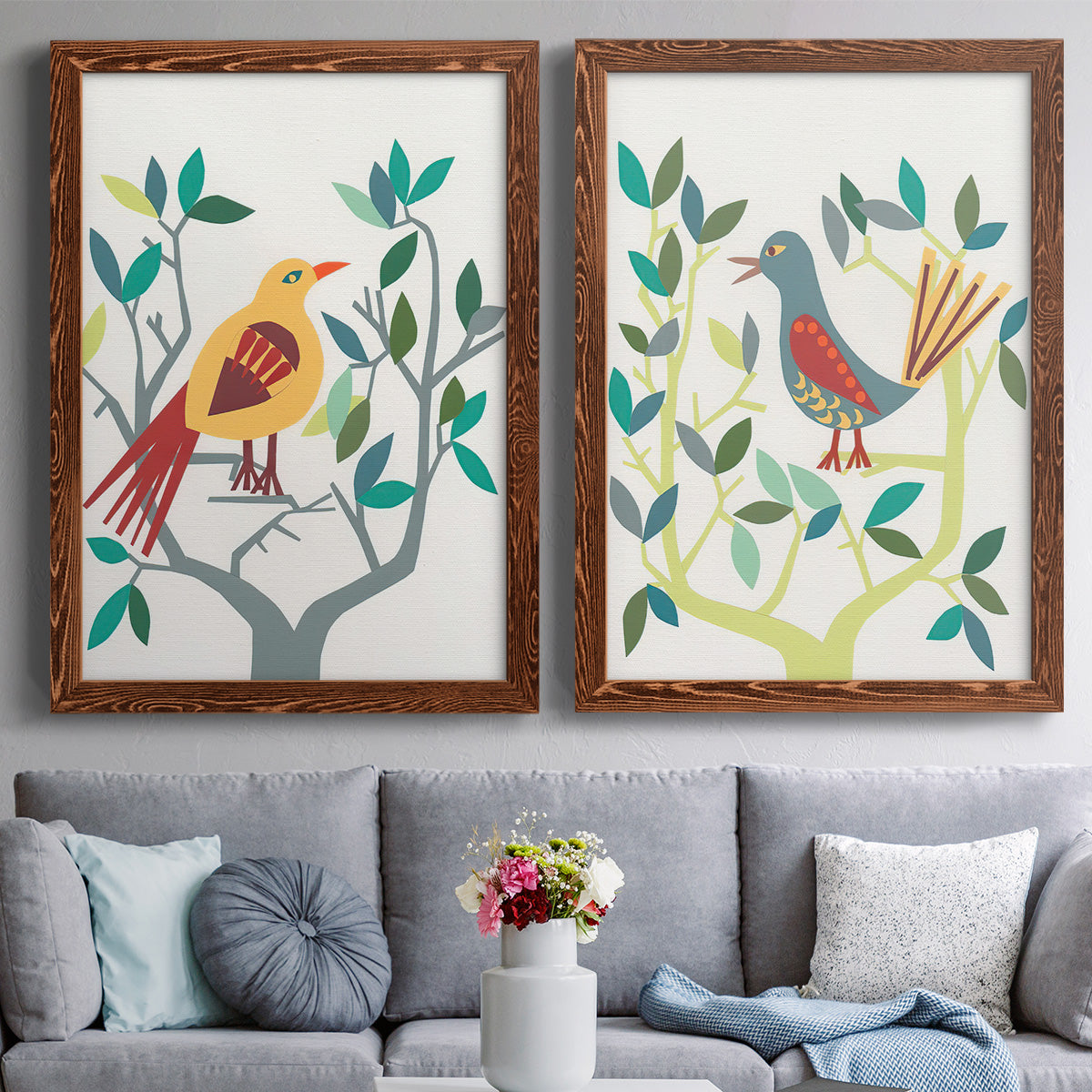 Set of two maple barnwood framed canvas prints depicting paired modern folk-art bird illustrations with red yellow blue plumage, simplified leaves, and playful flat shapes above a sofa in a contemporary living room.