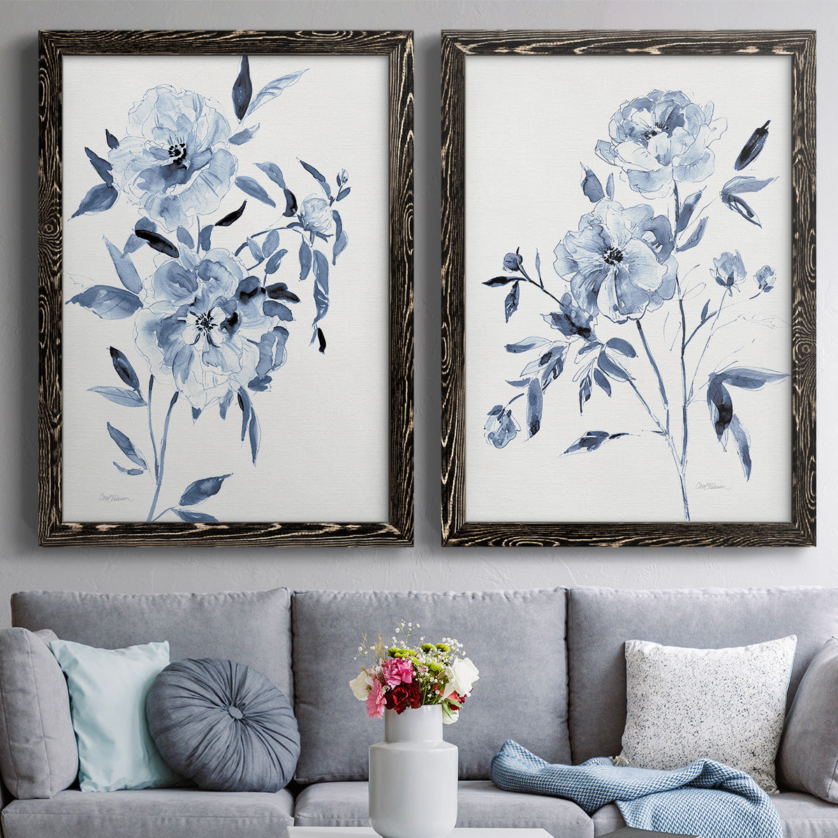 Set of two black barnwood framed canvas prints depicting paired expressive watercolor botanical paintings with indigo blossoms, gestural blue leaves, and airy white space above a sofa in a contemporary living room.