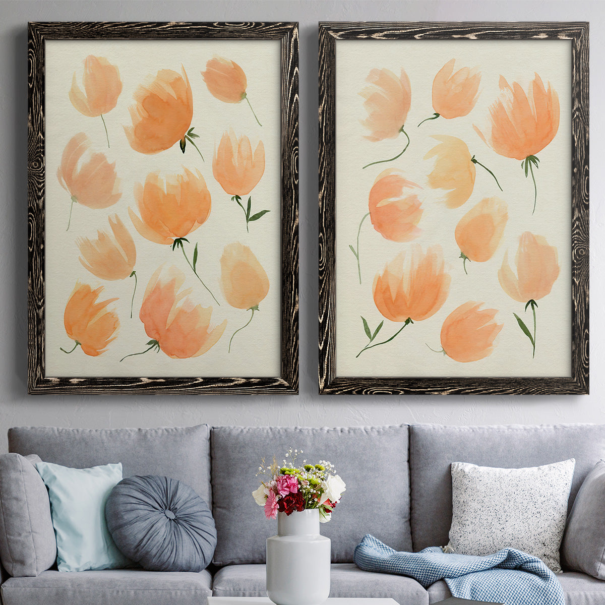 Set of two black barnwood framed canvas prints depicting paired airy watercolor floral paintings with soft petals, thin black stems, and warm cream backgrounds above a sofa in a contemporary living room.