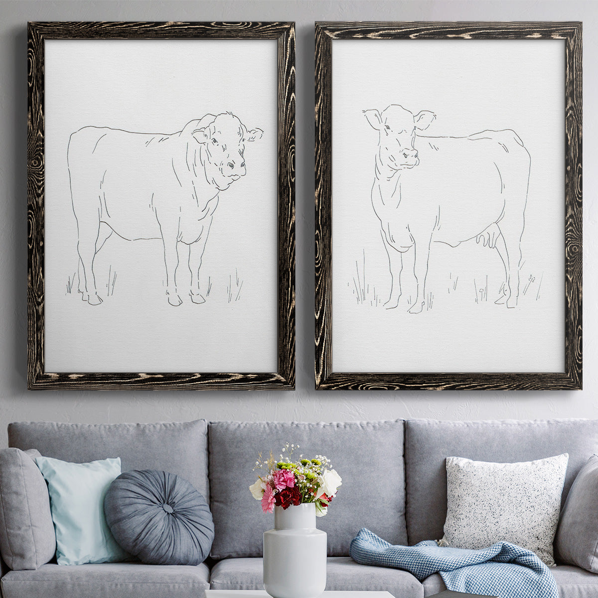 Set of two black barnwood framed canvas prints depicting paired minimalist livestock sketches with fine gray lines, white backgrounds, and quiet farm-animal profiles above a sofa in a contemporary living room.