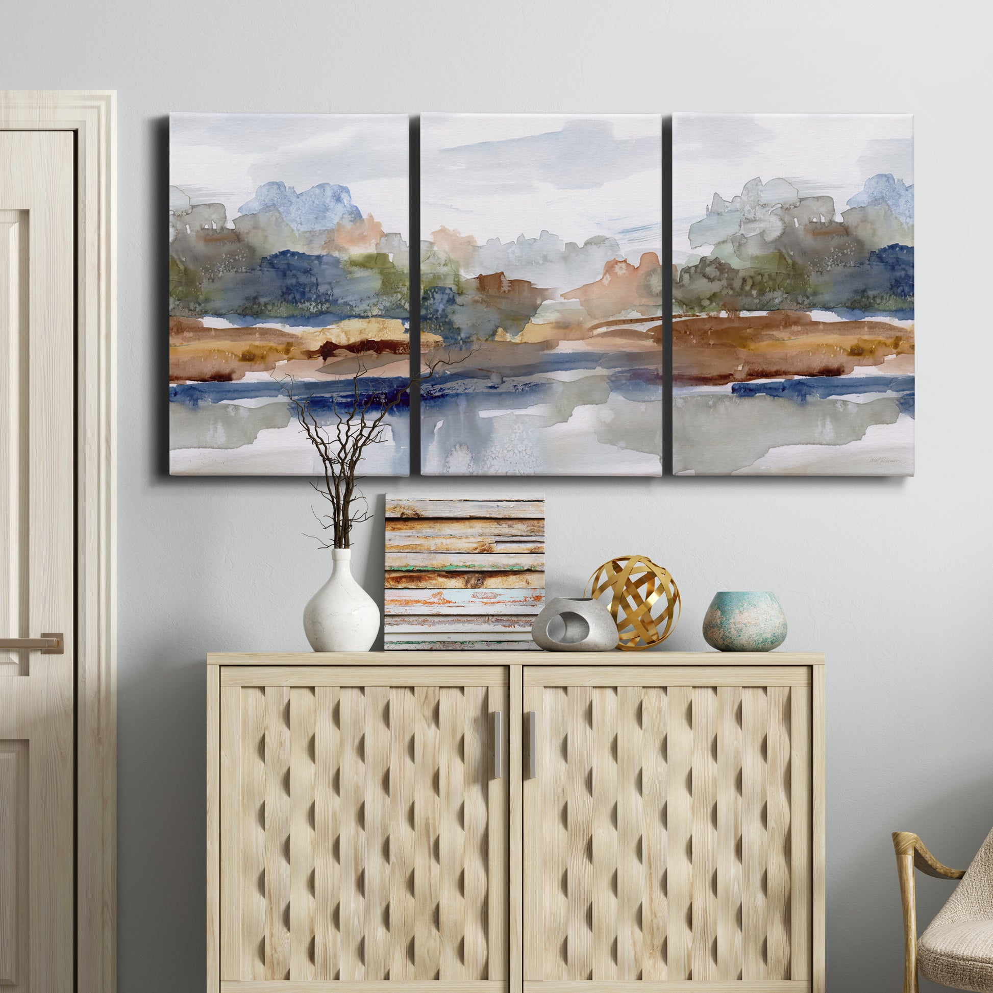 Three-panel canvas wall art featuring layered abstract mountain forms, displayed above a cabinet in an entryway.
