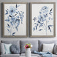 Set of two gray barnwood framed canvas prints depicting paired expressive watercolor botanical paintings with indigo blossoms, gestural blue leaves, and airy white space above a sofa in a contemporary living room.