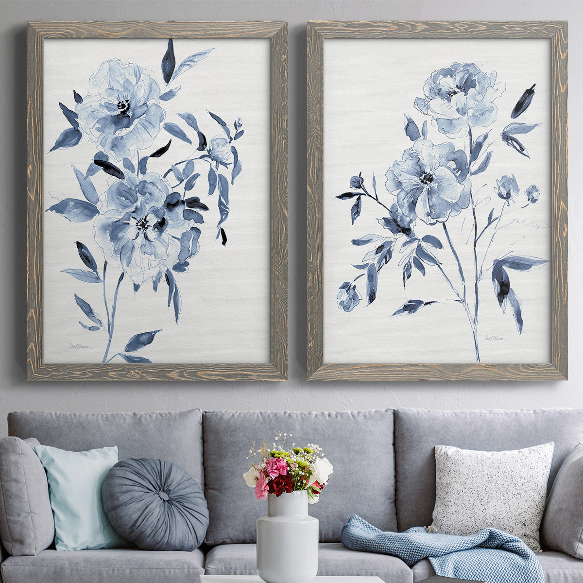 Set of two gray barnwood framed canvas prints depicting paired expressive watercolor botanical paintings with indigo blossoms, gestural blue leaves, and airy white space above a sofa in a contemporary living room.