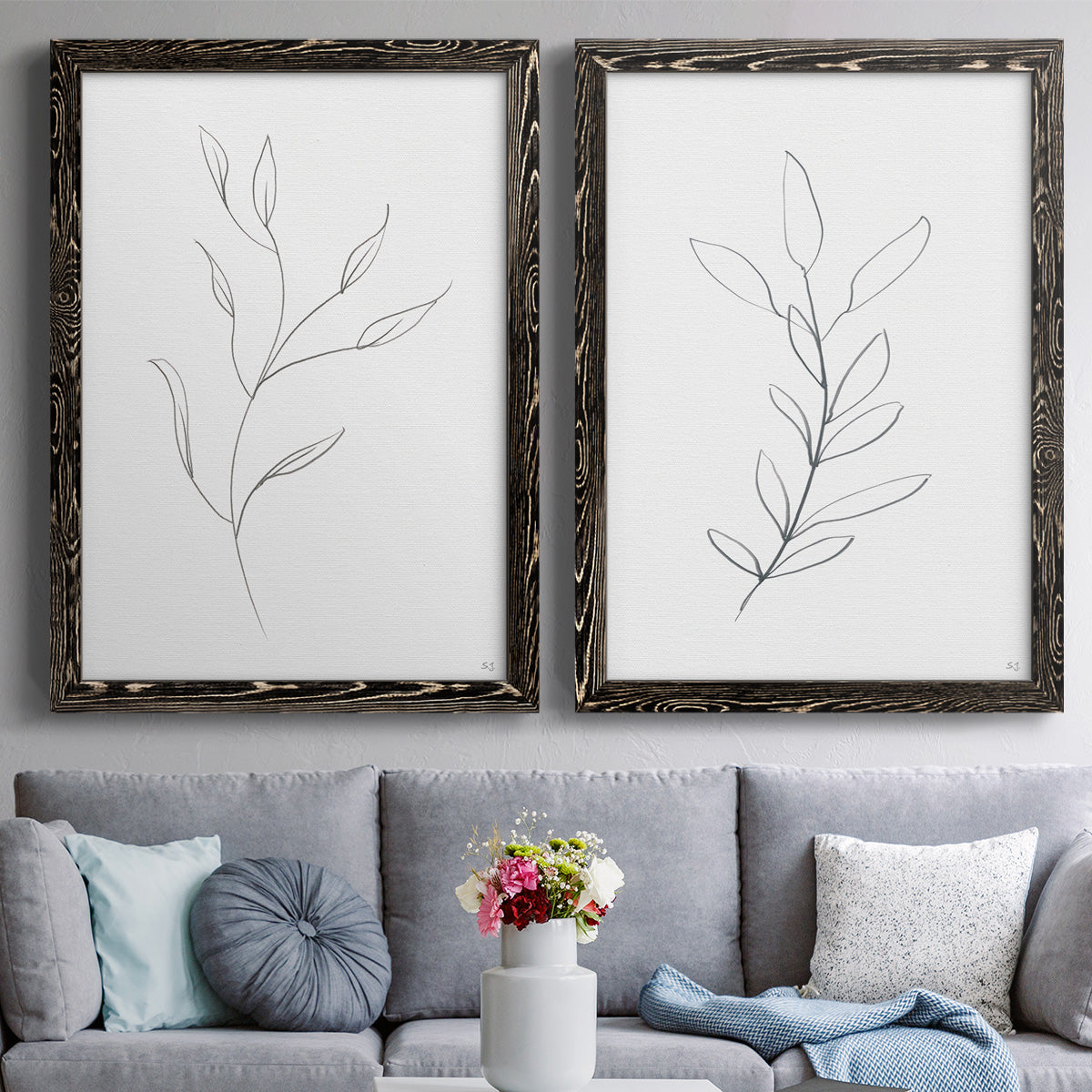 Set of two black barnwood framed canvas prints depicting paired minimalist botanical line drawings with thin gray contours, sparse stems, and clean white fields above a sofa in a contemporary living room.