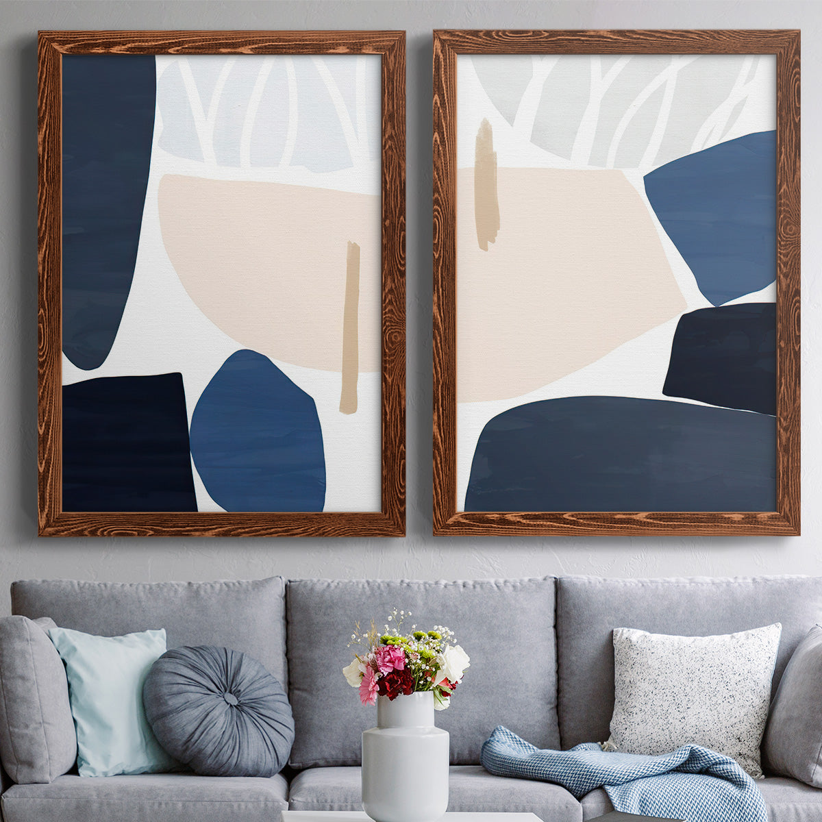Set of two maple barnwood framed canvas prints depicting paired minimalist color-block abstracts with rounded dark forms, pale sand fields, and clean modern edges above a sofa in a contemporary living room.