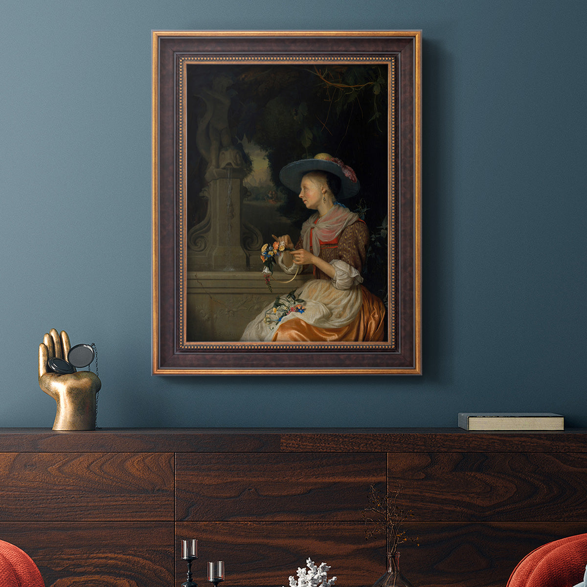 Classical interior scene of a woman weaving flowers by Godfried Schalcken in an ornate bronzed mahogany wood frame displayed in a dark-toned interior setting.
