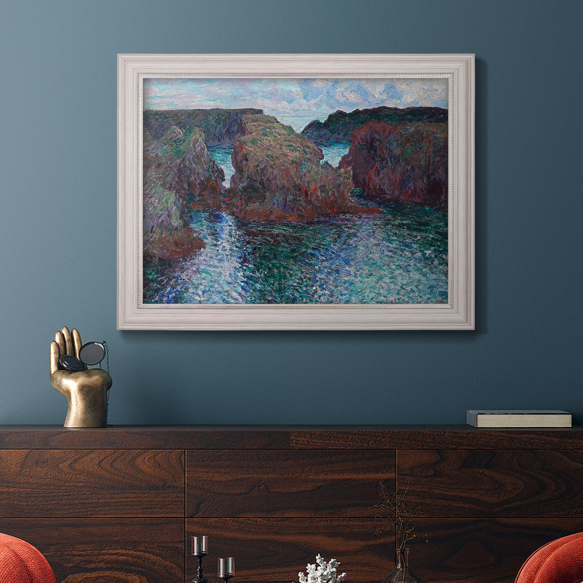 Impressionist rocky seascape by Claude Monet in an ornate weathered white frame displayed in a dark-toned interior setting.