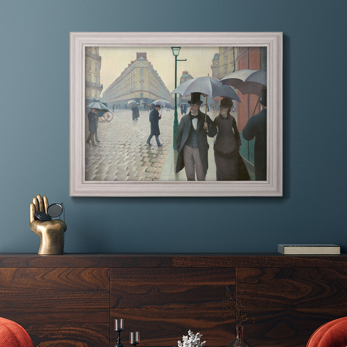 Impressionist urban street scene in the rain by Gustave Caillebotte in an ornate weathered white frame displayed in a dark-toned interior setting.