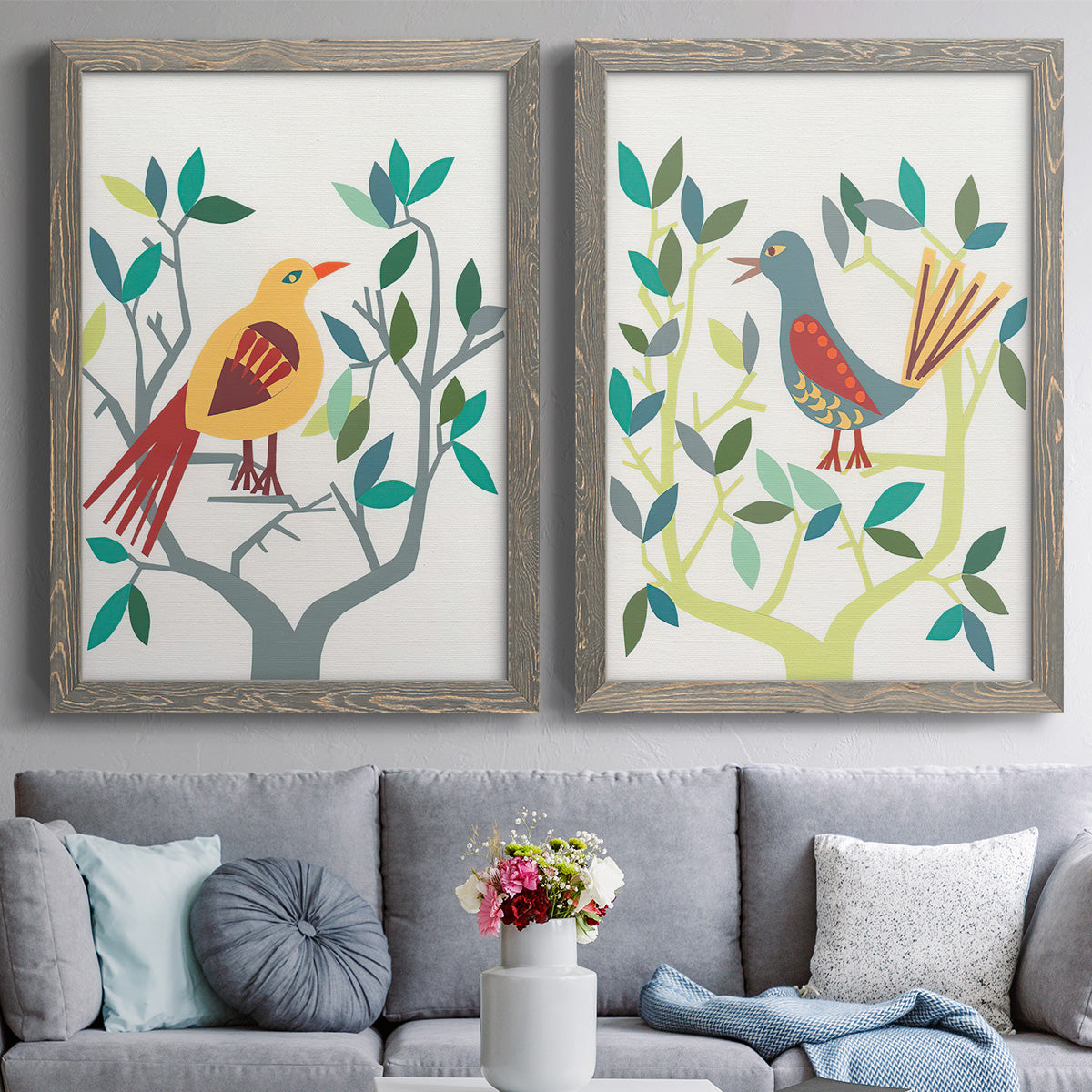 Set of two gray barnwood framed canvas prints depicting paired modern folk-art bird illustrations with red yellow blue plumage, simplified leaves, and playful flat shapes above a sofa in a contemporary living room.