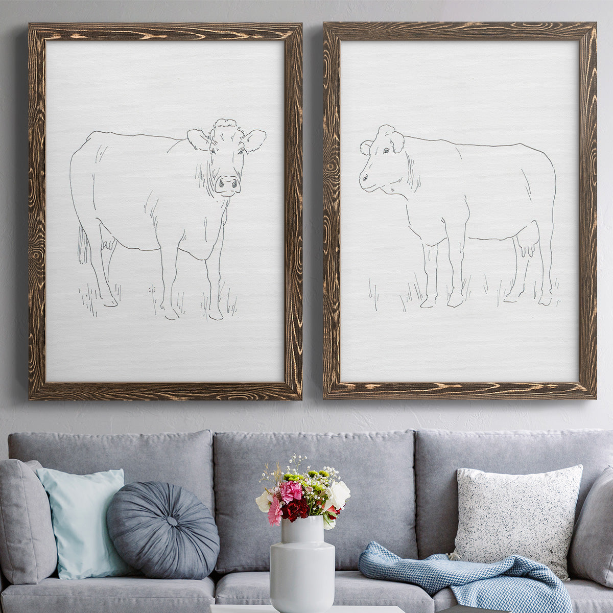 Set of two walnut barnwood framed canvas prints depicting paired minimalist livestock sketches with fine gray lines, white backgrounds, and quiet farm-animal profiles above a sofa in a contemporary living room.