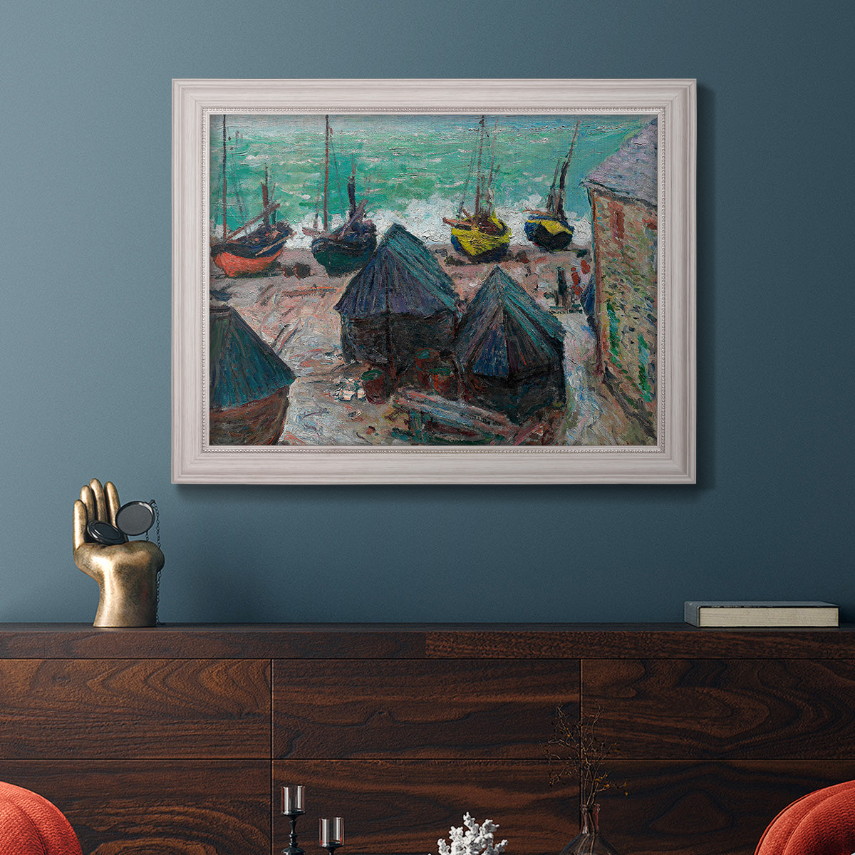 Impressionist coastal beach scene with boats by Claude Monet in an ornate weathered white frame displayed in a dark-toned interior setting.