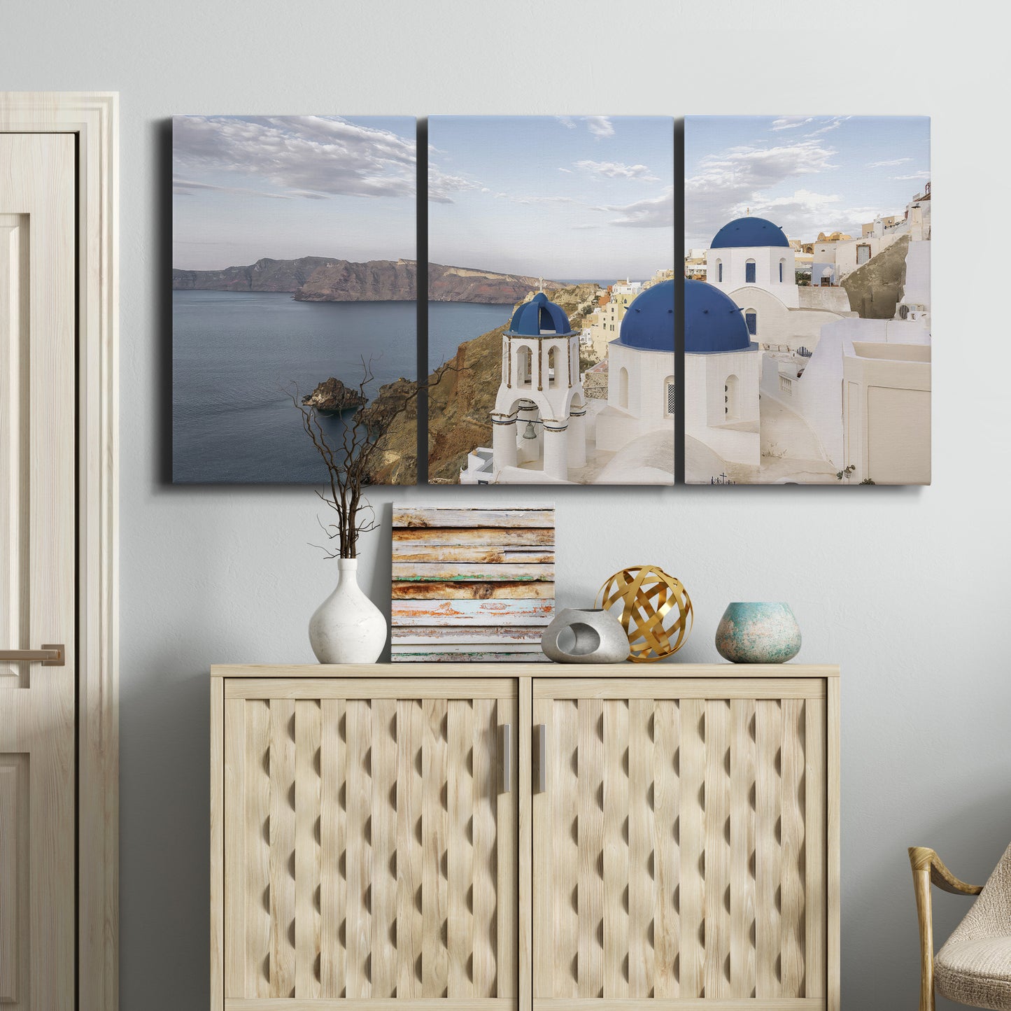 Three-panel canvas wall art featuring white buildings overlooking sea cliffs, displayed above a cabinet in an entryway.
