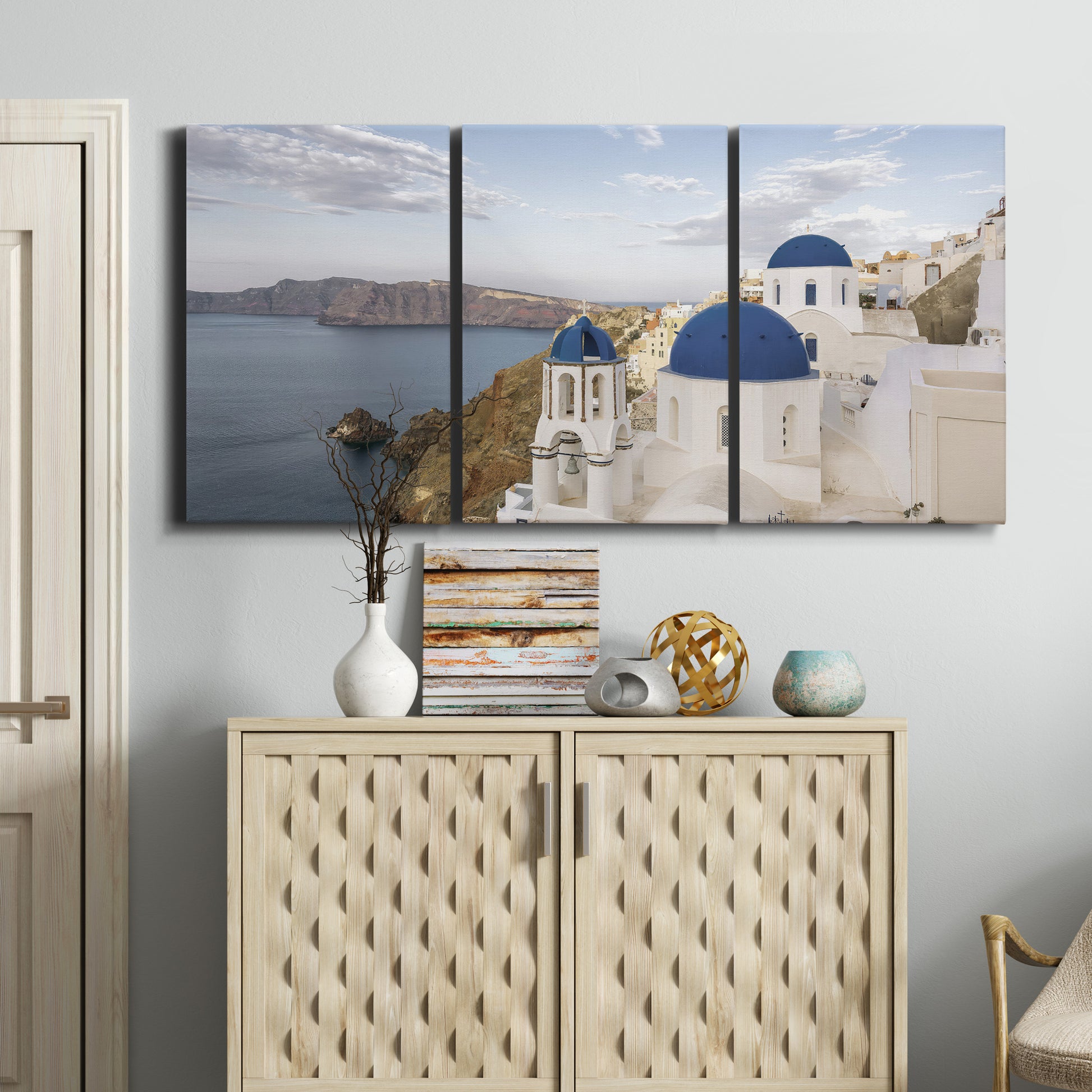 Three-panel canvas wall art featuring white buildings overlooking sea cliffs, displayed above a cabinet in an entryway.