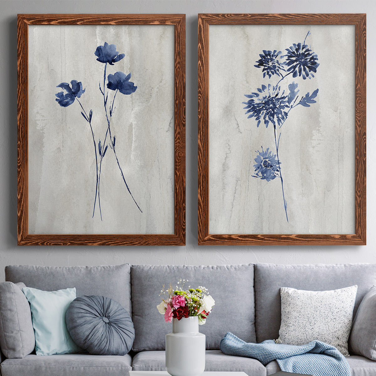 Set of two maple barnwood framed canvas prints depicting paired rustic monochrome botanical paintings with indigo blossoms, thin stems, and softly distressed gray backgrounds above a sofa in a contemporary living room.