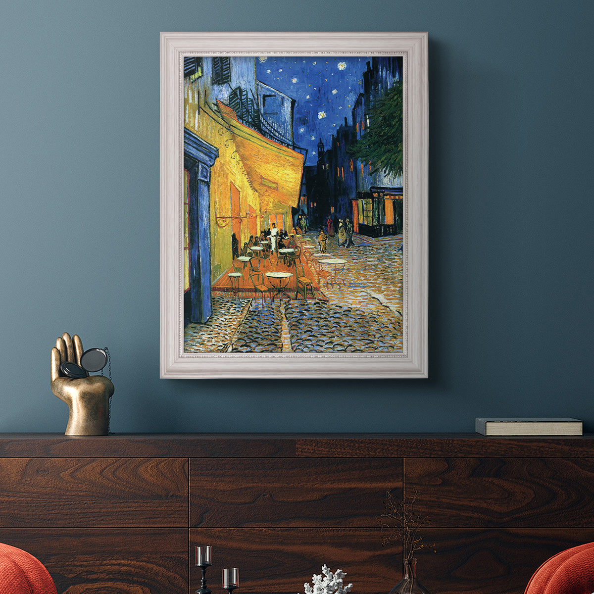 Impressionist night café street scene by Vincent van Gogh in an ornate weathered white frame displayed in a dark-toned interior setting.