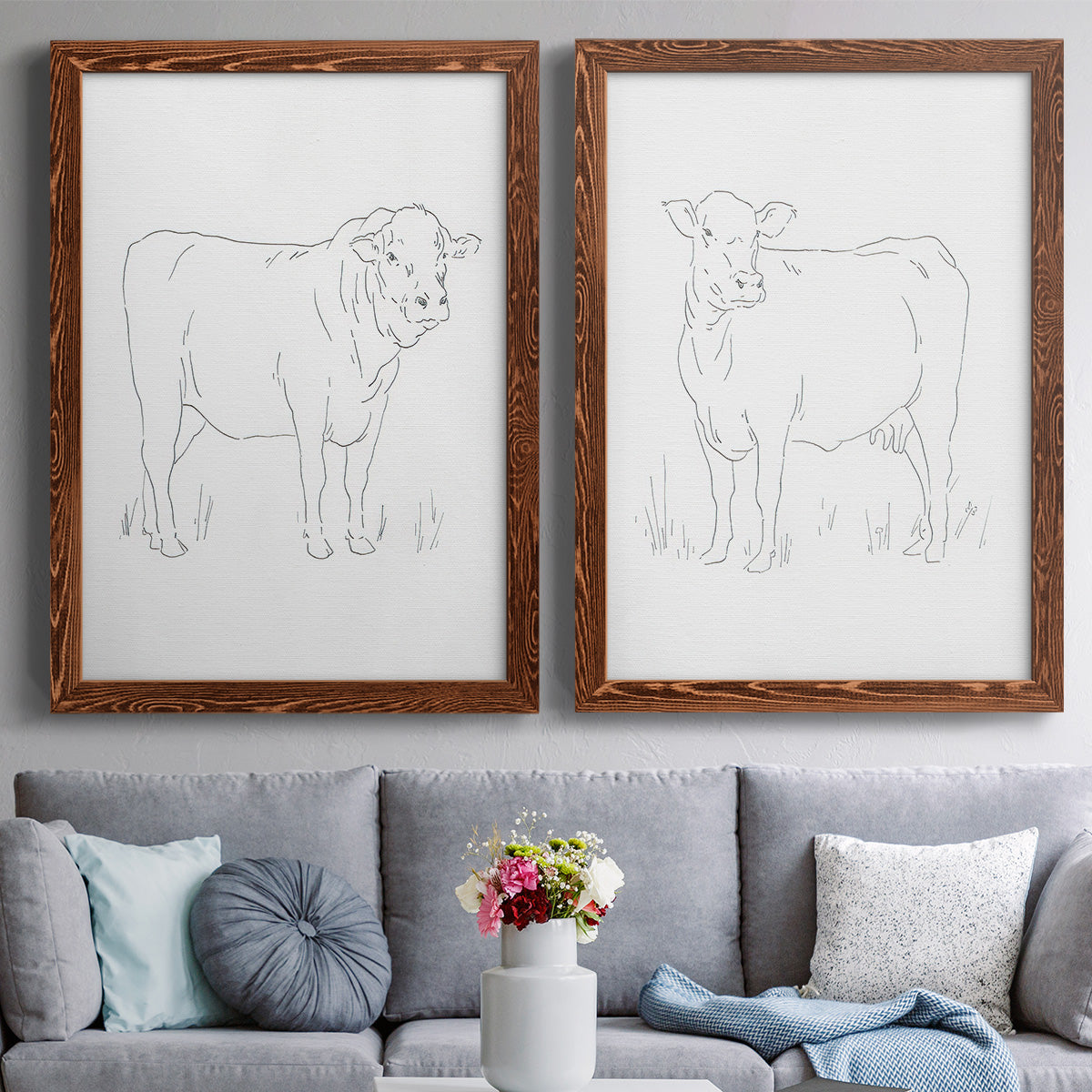 Set of two maple barnwood framed canvas prints depicting paired minimalist livestock sketches with fine gray lines, white backgrounds, and quiet farm-animal profiles above a sofa in a contemporary living room.