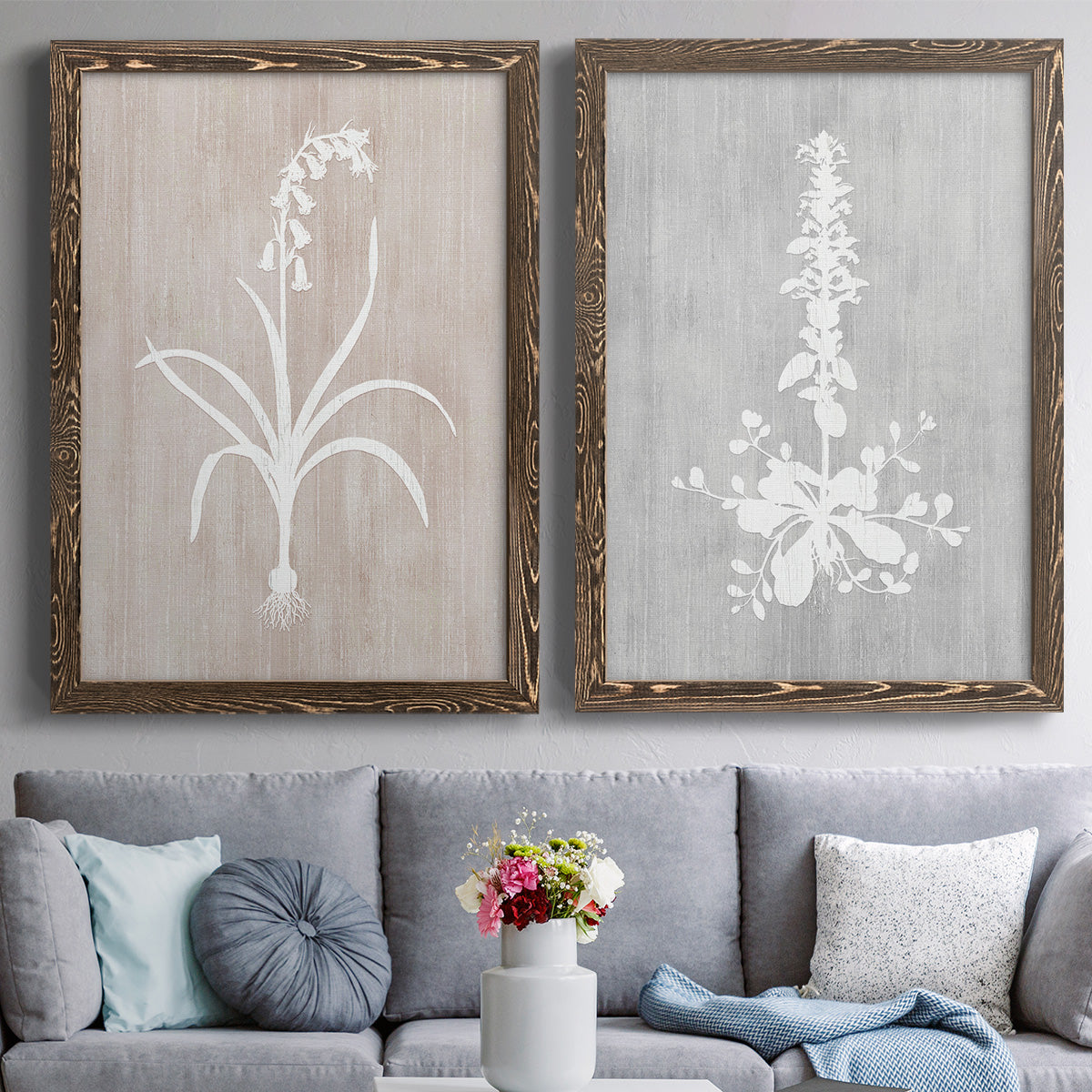 Set of two walnut barnwood framed canvas prints depicting paired rustic minimalist botanical prints with delicate stems, leaves, and blossoms against gray taupe and pale blue panels above a sofa in a contemporary living room.