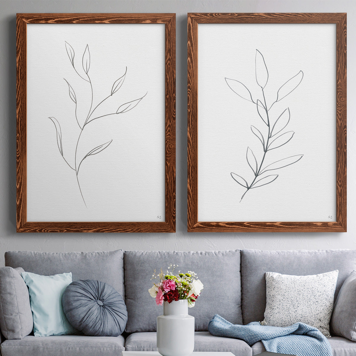 Set of two maple barnwood framed canvas prints depicting paired minimalist botanical line drawings with thin gray contours, sparse stems, and clean white fields above a sofa in a contemporary living room.