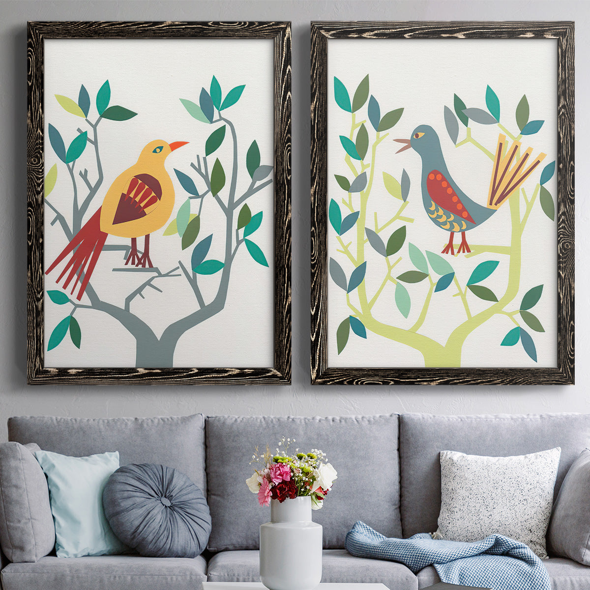 Set of two black barnwood framed canvas prints depicting paired modern folk-art bird illustrations with red yellow blue plumage, simplified leaves, and playful flat shapes above a sofa in a contemporary living room.