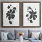 Set of two walnut barnwood framed canvas prints depicting paired minimalist botanical prints with bold dark leaves, curving stems, and clean white grounds above a sofa in a contemporary living room.