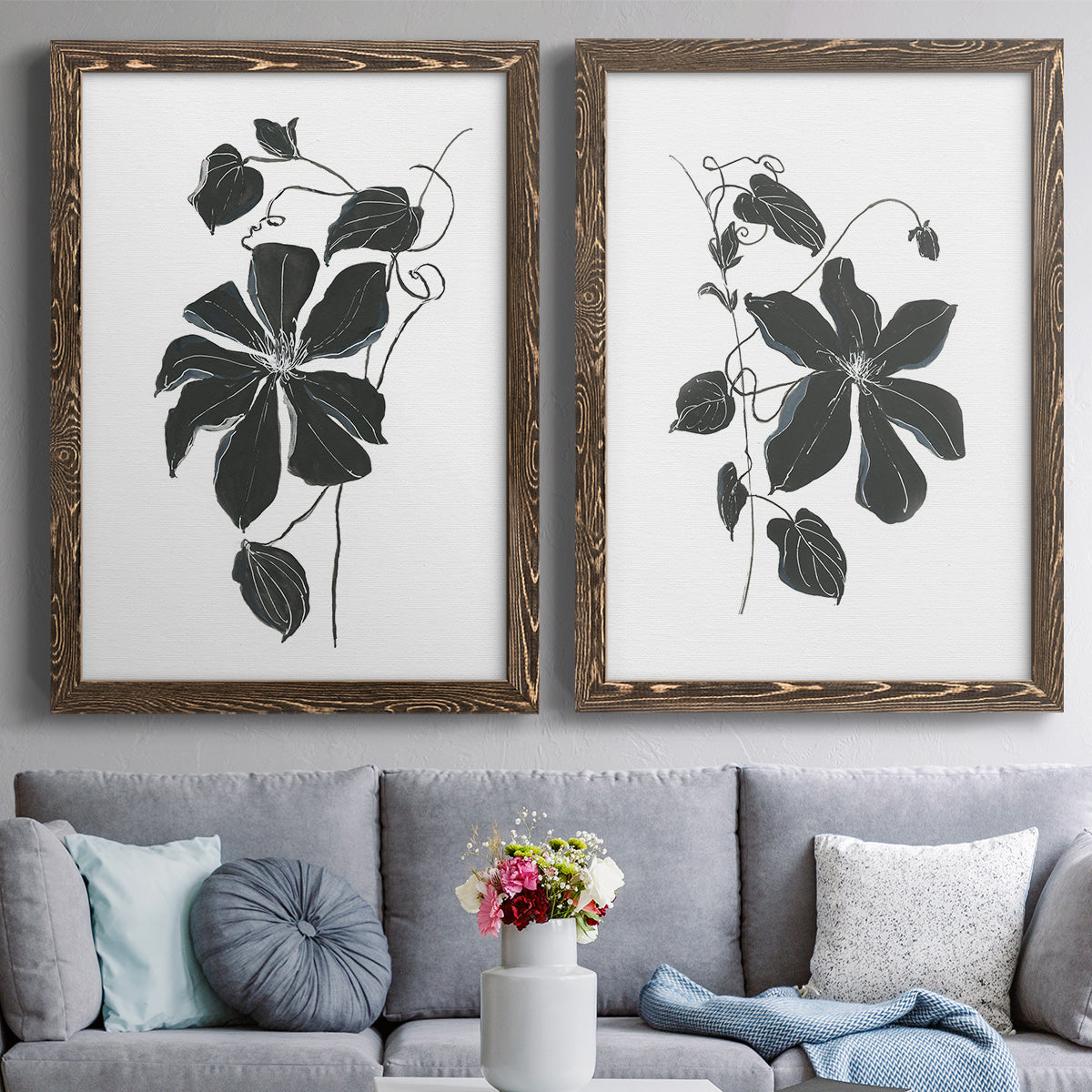 Set of two walnut barnwood framed canvas prints depicting paired minimalist botanical prints with bold dark leaves, curving stems, and clean white grounds above a sofa in a contemporary living room.