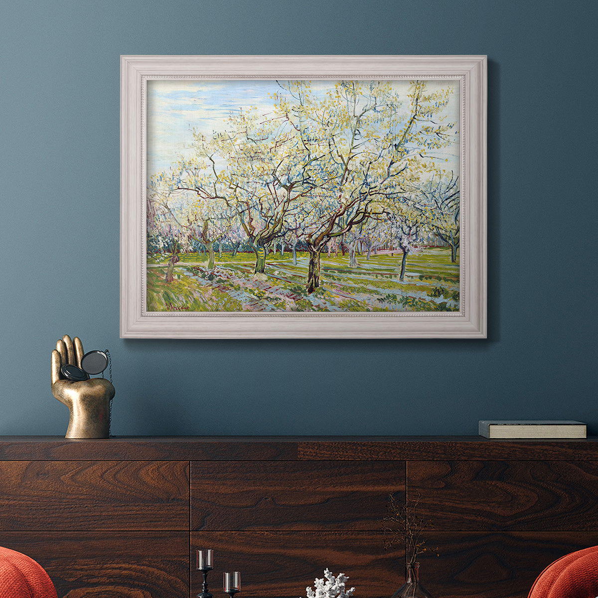 Impressionist white-blossomed orchard landscape by Vincent van Gogh in an ornate weathered white frame displayed in a dark-toned interior setting.