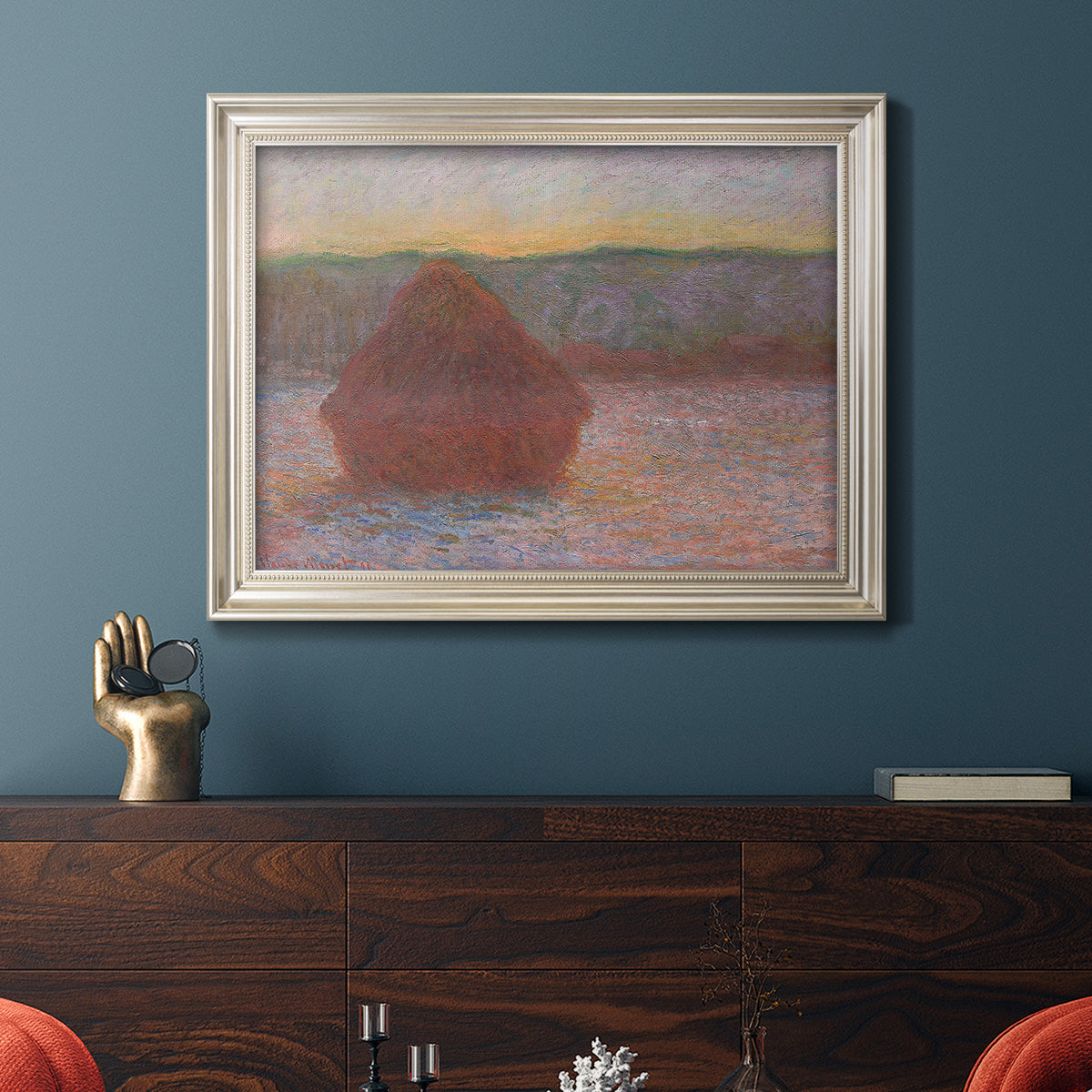 Impressionist haystack sunset scene in winter by Claude Monet in an ornate champagne frame displayed in a dark-toned interior setting.