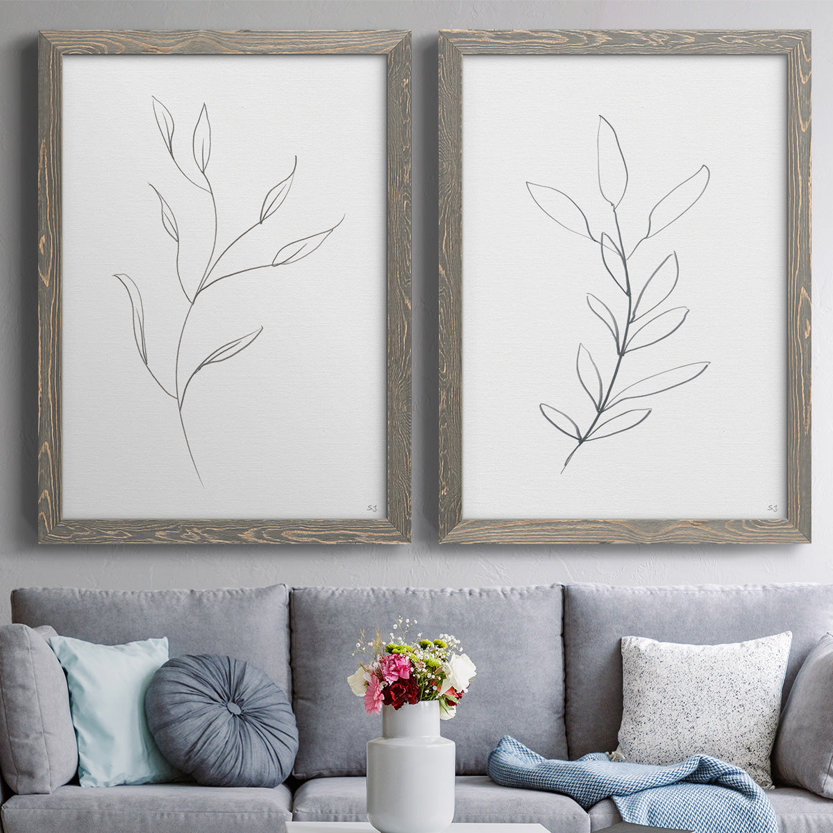 Set of two gray barnwood framed canvas prints depicting paired minimalist botanical line drawings with thin gray contours, sparse stems, and clean white fields above a sofa in a contemporary living room.