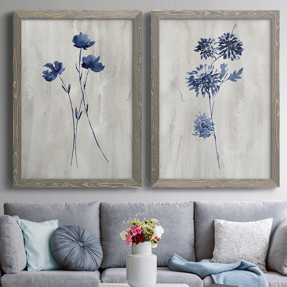 Set of two gray barnwood framed canvas prints depicting paired rustic monochrome botanical paintings with indigo blossoms, thin stems, and softly distressed gray backgrounds above a sofa in a contemporary living room.