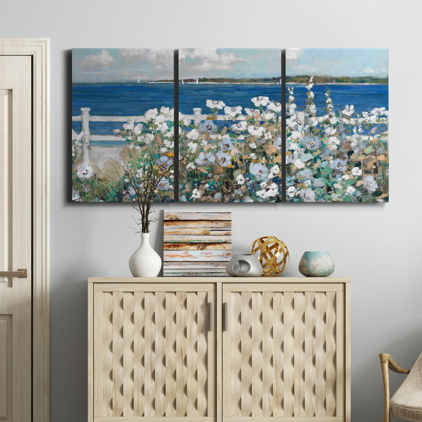 Three-panel canvas wall art featuring coastal garden overlooking bay, displayed above a cabinet in an entryway.