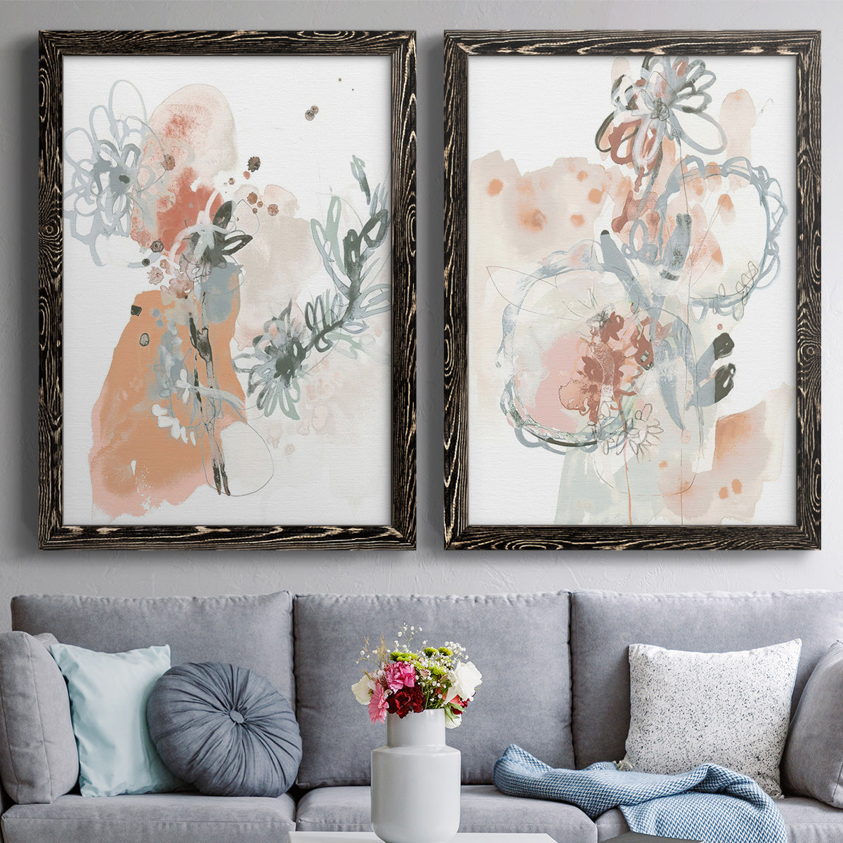 Set of two black barnwood framed canvas prints depicting paired abstract watercolor botanicals with scribbled petals, translucent washes, and drifting stems above a sofa in a contemporary living room.