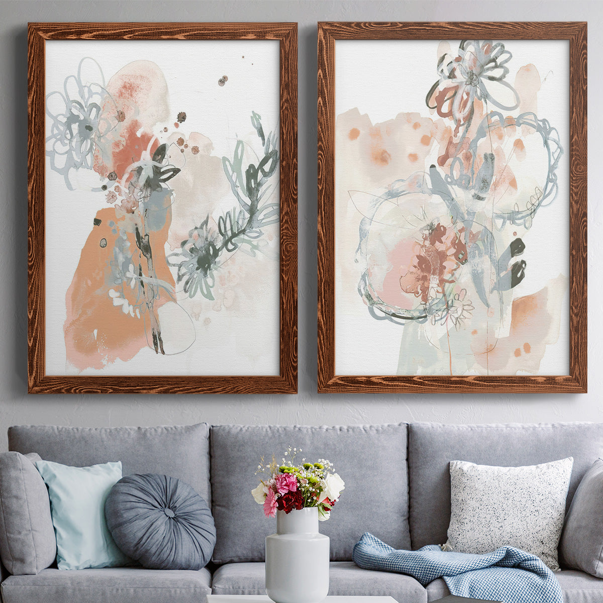 Set of two maple barnwood framed canvas prints depicting paired abstract watercolor botanicals with scribbled petals, translucent washes, and drifting stems above a sofa in a contemporary living room.
