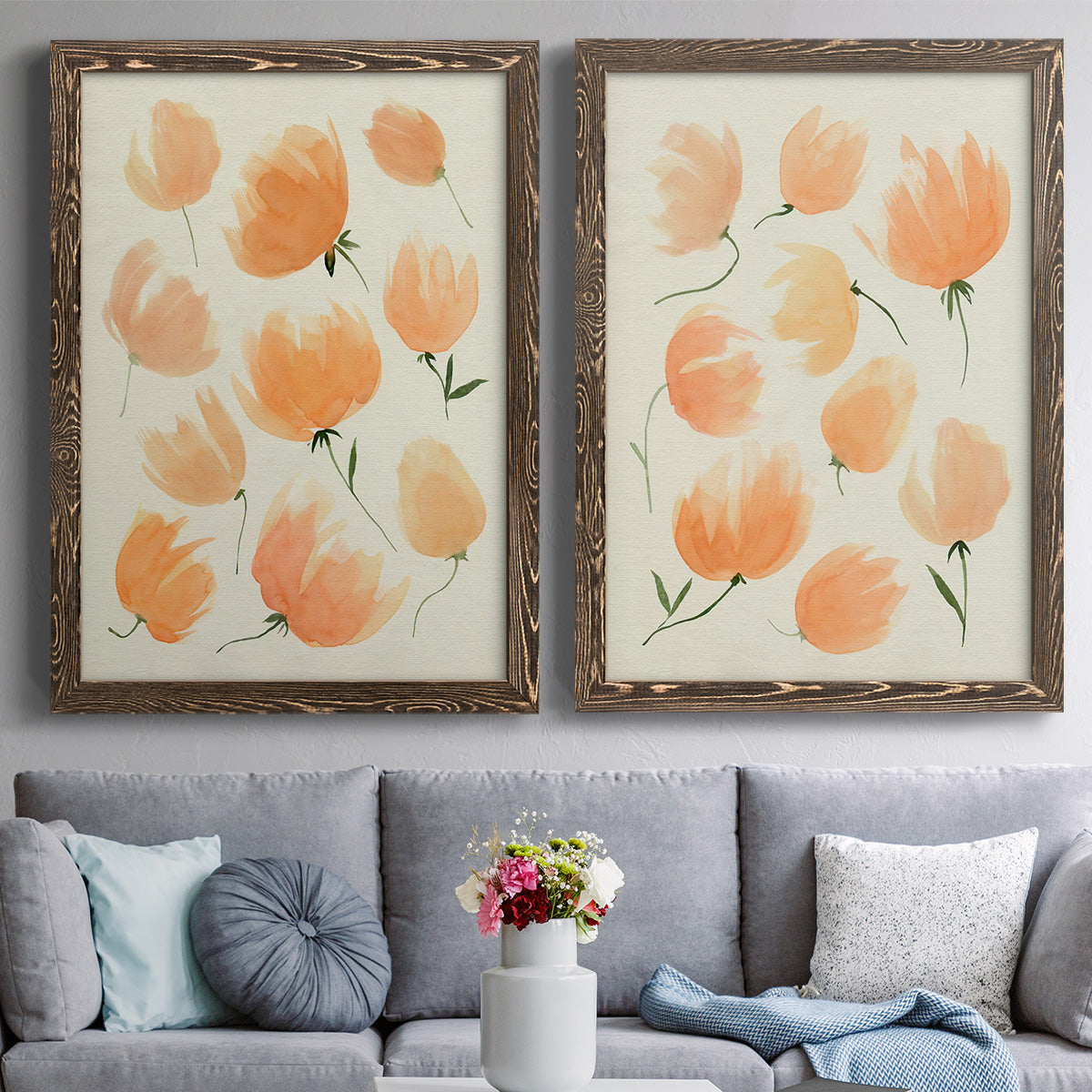 Set of two walnut barnwood framed canvas prints depicting paired airy watercolor floral paintings with soft petals, thin black stems, and warm cream backgrounds above a sofa in a contemporary living room.