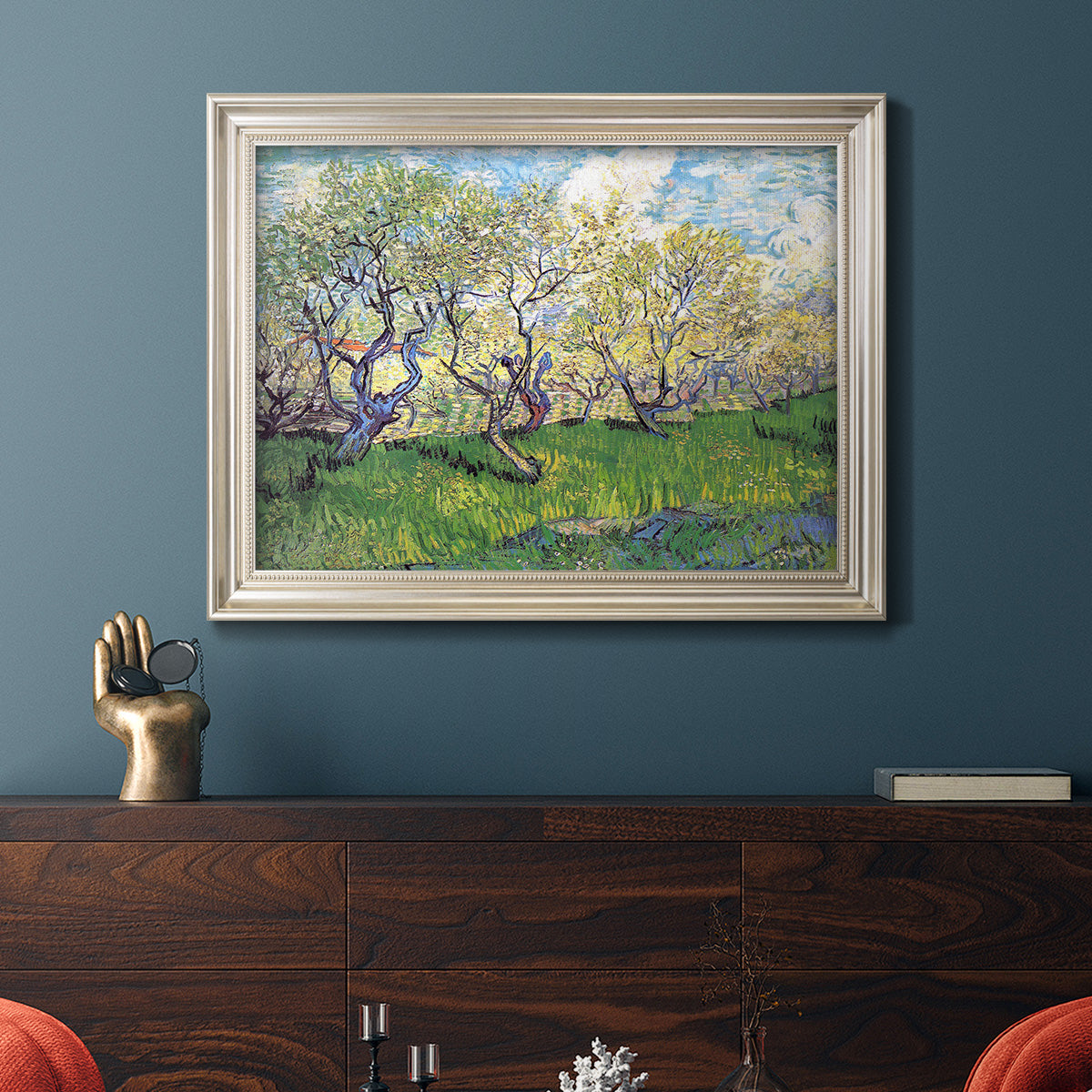 Impressionist spring orchard landscape by Vincent van Gogh in an ornate champagne frame displayed in a dark-toned interior setting.