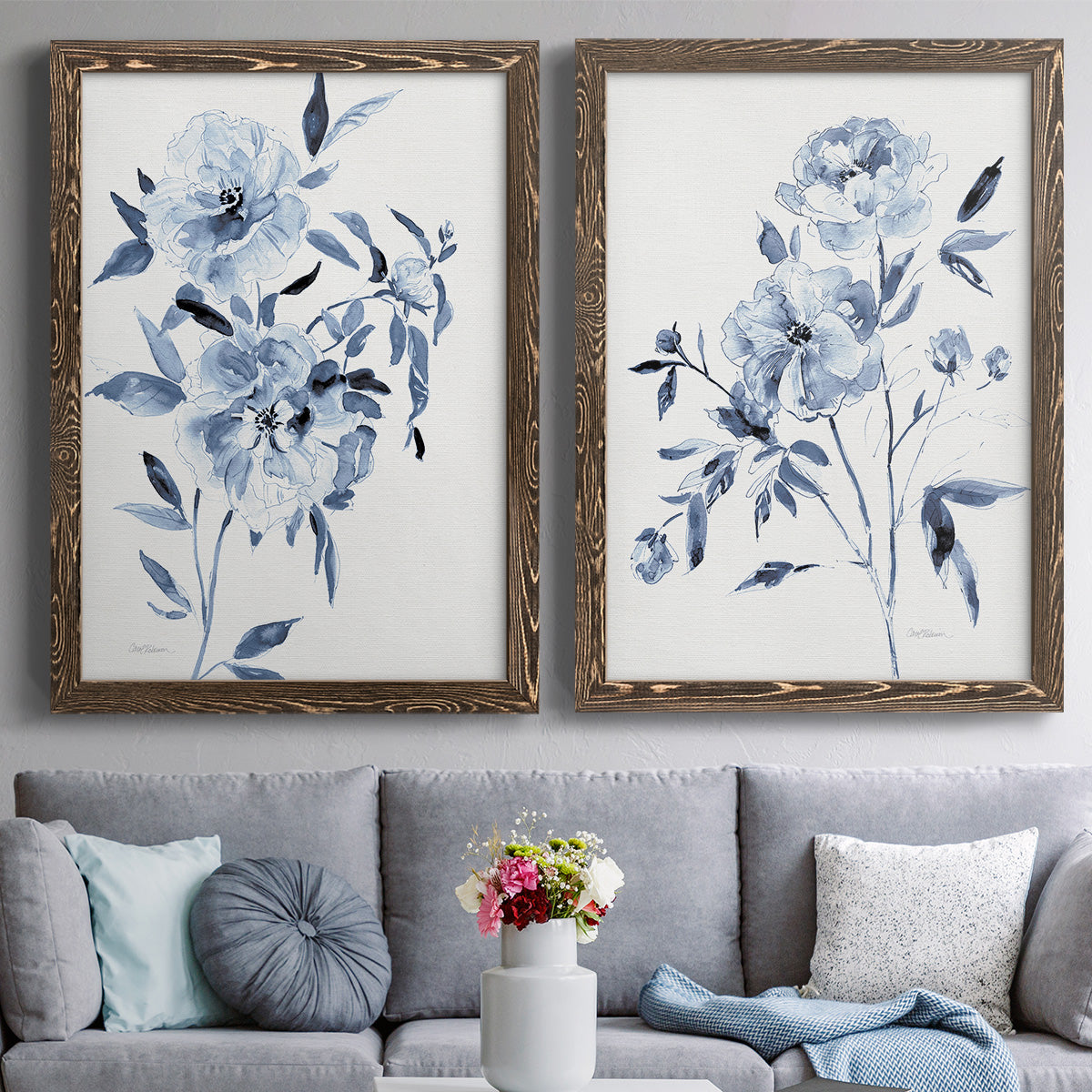 Set of two walnut barnwood framed canvas prints depicting paired expressive watercolor botanical paintings with indigo blossoms, gestural blue leaves, and airy white space above a sofa in a contemporary living room.