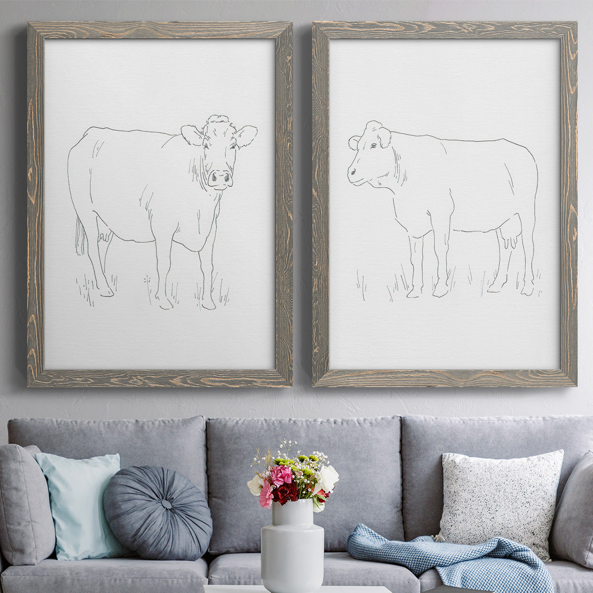 Set of two gray barnwood framed canvas prints depicting paired minimalist livestock sketches with fine gray lines, white backgrounds, and quiet farm-animal profiles above a sofa in a contemporary living room.