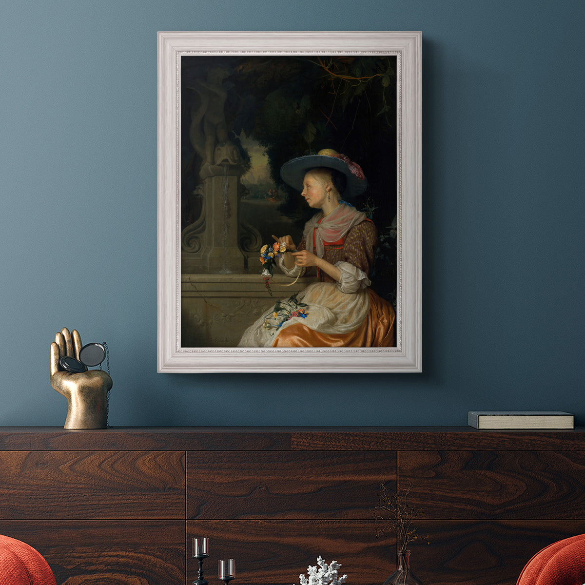 Classical interior scene of a woman weaving flowers by Godfried Schalcken in an ornate weathered white frame displayed in a dark-toned interior setting.