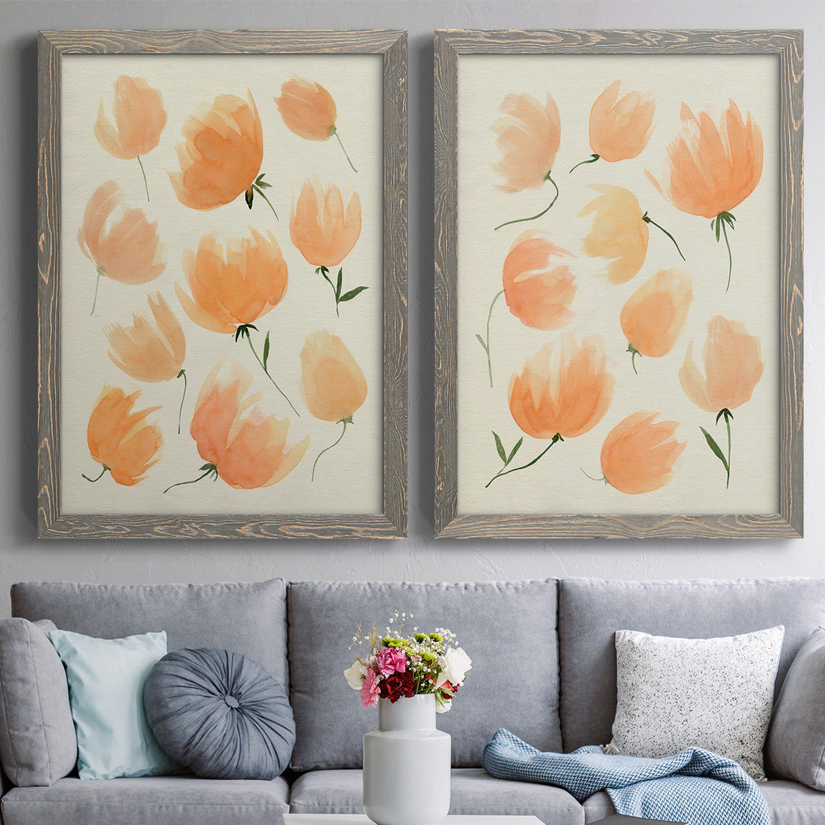 Set of two gray barnwood framed canvas prints depicting paired airy watercolor floral paintings with soft petals, thin black stems, and warm cream backgrounds above a sofa in a contemporary living room.