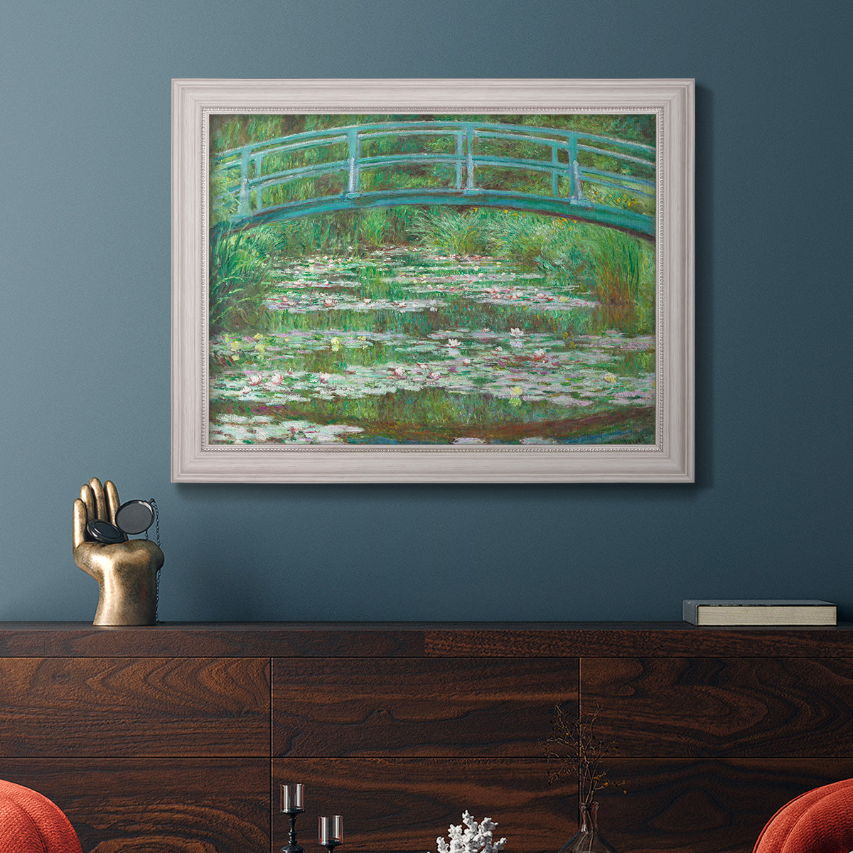 Impressionist garden scene with Japanese bridge and water lilies by Claude Monet in an ornate weathered white frame displayed in a dark-toned interior setting.