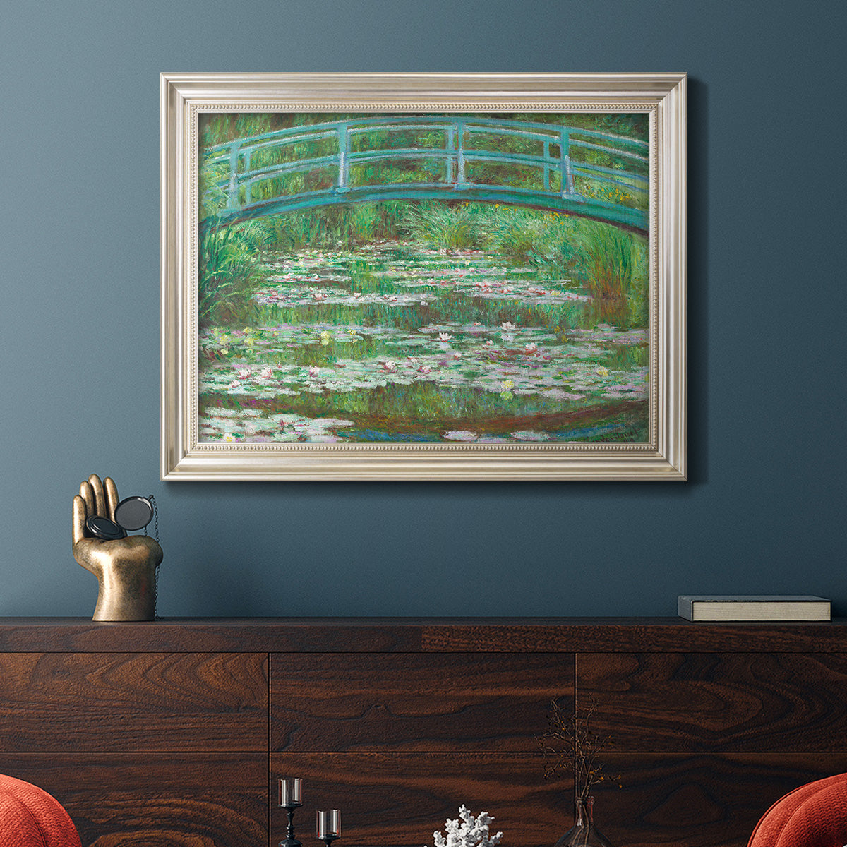 Impressionist garden scene with Japanese bridge and water lilies by Claude Monet in an ornate champagne frame displayed in a dark-toned interior setting.