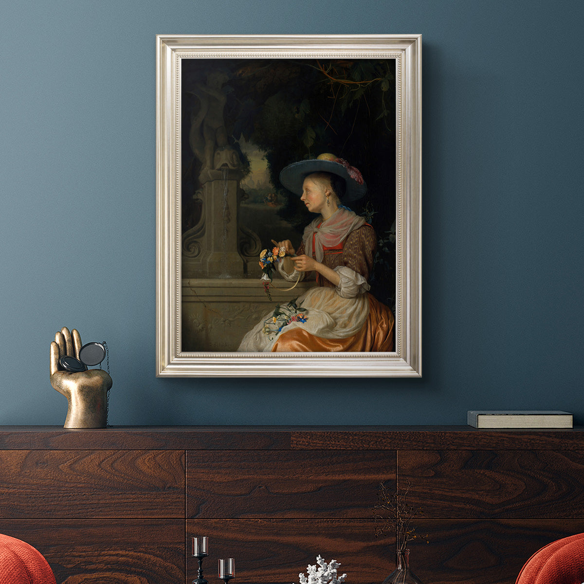 Classical interior scene of a woman weaving flowers by Godfried Schalcken in an ornate champagne frame displayed in a dark-toned interior setting.