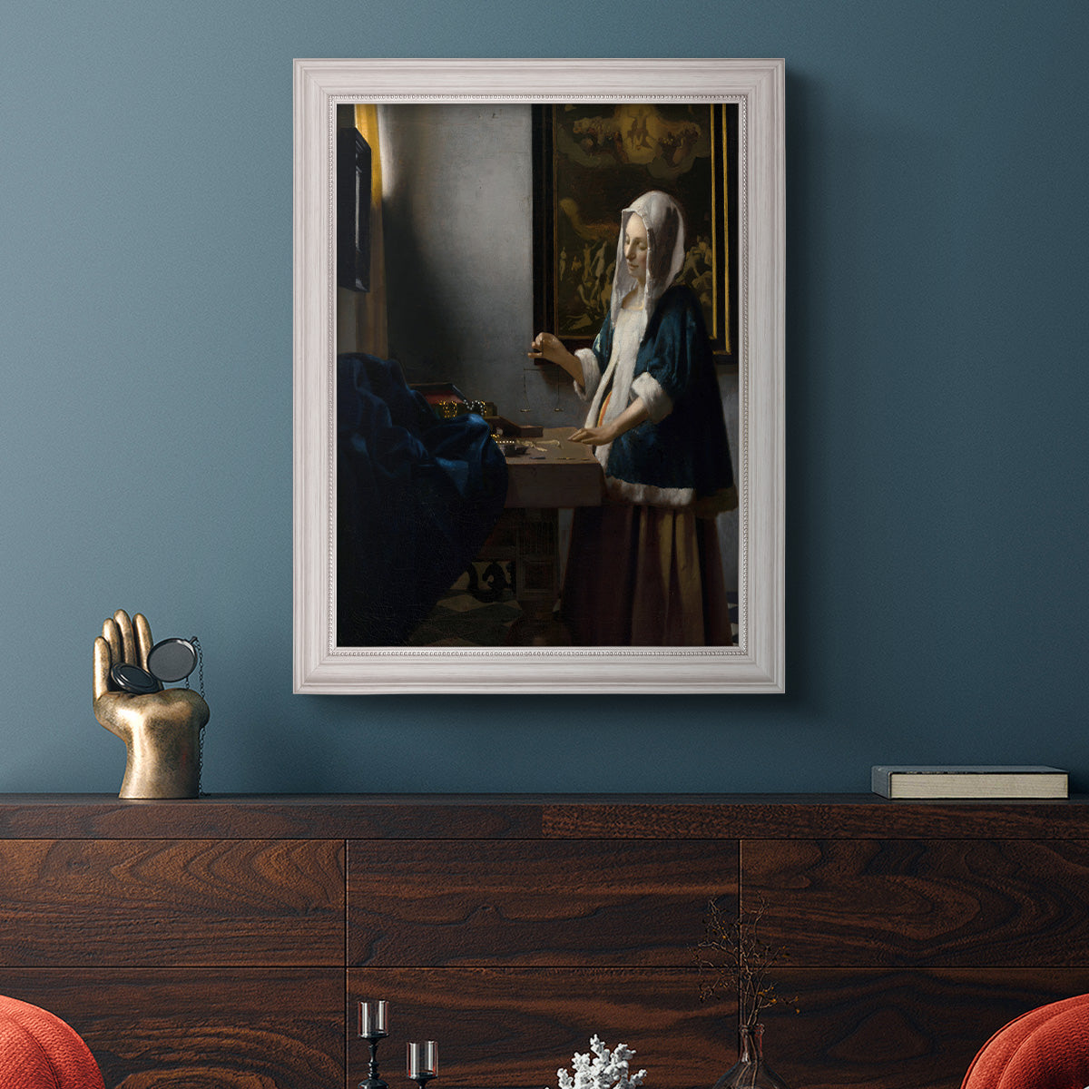 Quiet interior genre scene of a woman with scales by Johannes Vermeer in an ornate weathered white frame displayed in a dark-toned interior setting.