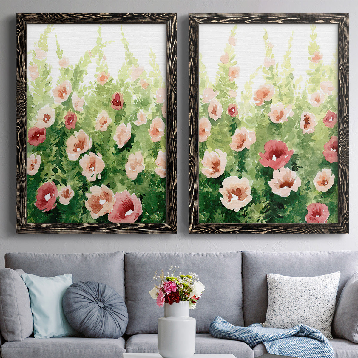 Set of two black barnwood framed canvas prints depicting paired luminous impressionistic floral paintings with loose blossoms, green stems, and sunlit watercolor texture above a sofa in a contemporary living room.