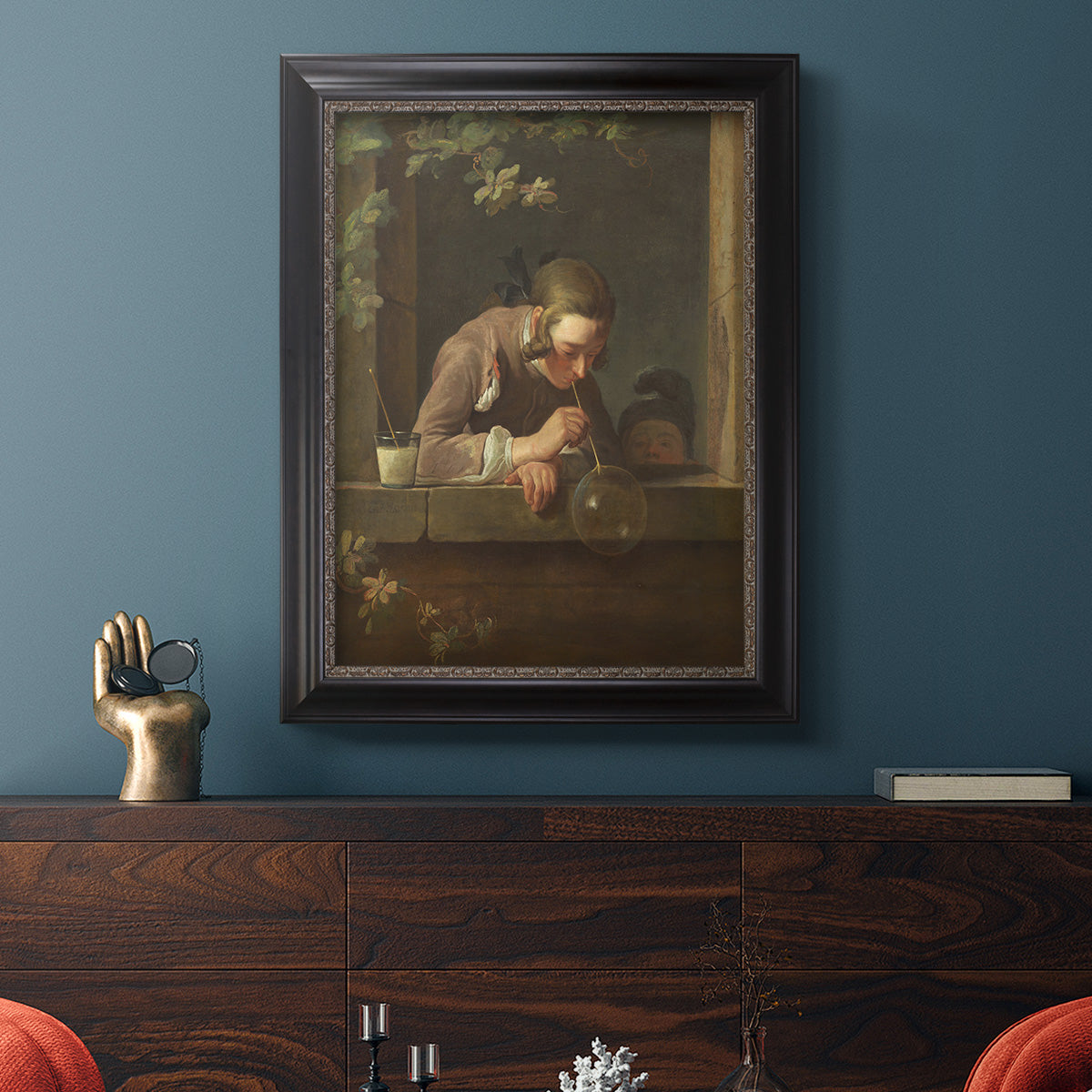 Classical genre painting of a child blowing soap bubbles by Jean-Siméon Chardin in an ornate black frame with gilded inner detail displayed in a dark-toned interior setting.